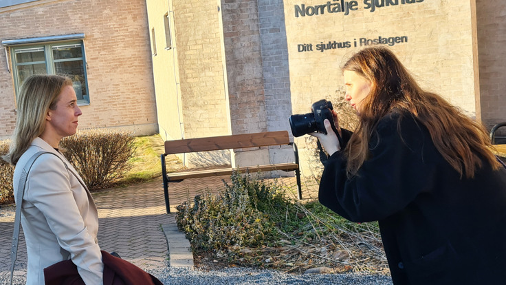 En kvinna fotograferar en annan kvinna framför en solbelyst vägg med texten Norrtälje sjukhus.