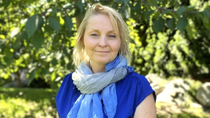 En kvinna i blå klänning står på gräsmatta, framför grönskande träd.