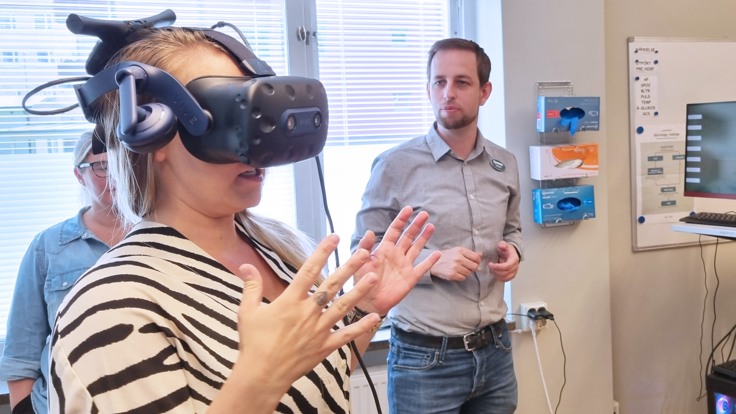 En kvinna med VR-glasögon på sig gestikulerar. En man står i bakgrunden och iakttar henne.