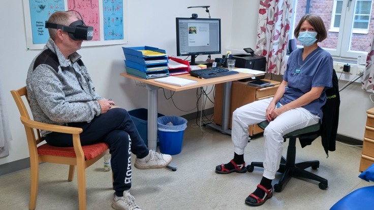 En man sitter på en stol med  VR-glasögon .  En kvinna med sjukhuskläder och munskydd sitter bredvid. 