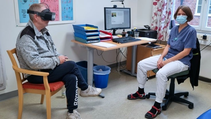 En man sitter på en stol med  VR-glasögon .  En kvinna med sjukhuskläder och munskydd sitter bredvid. 