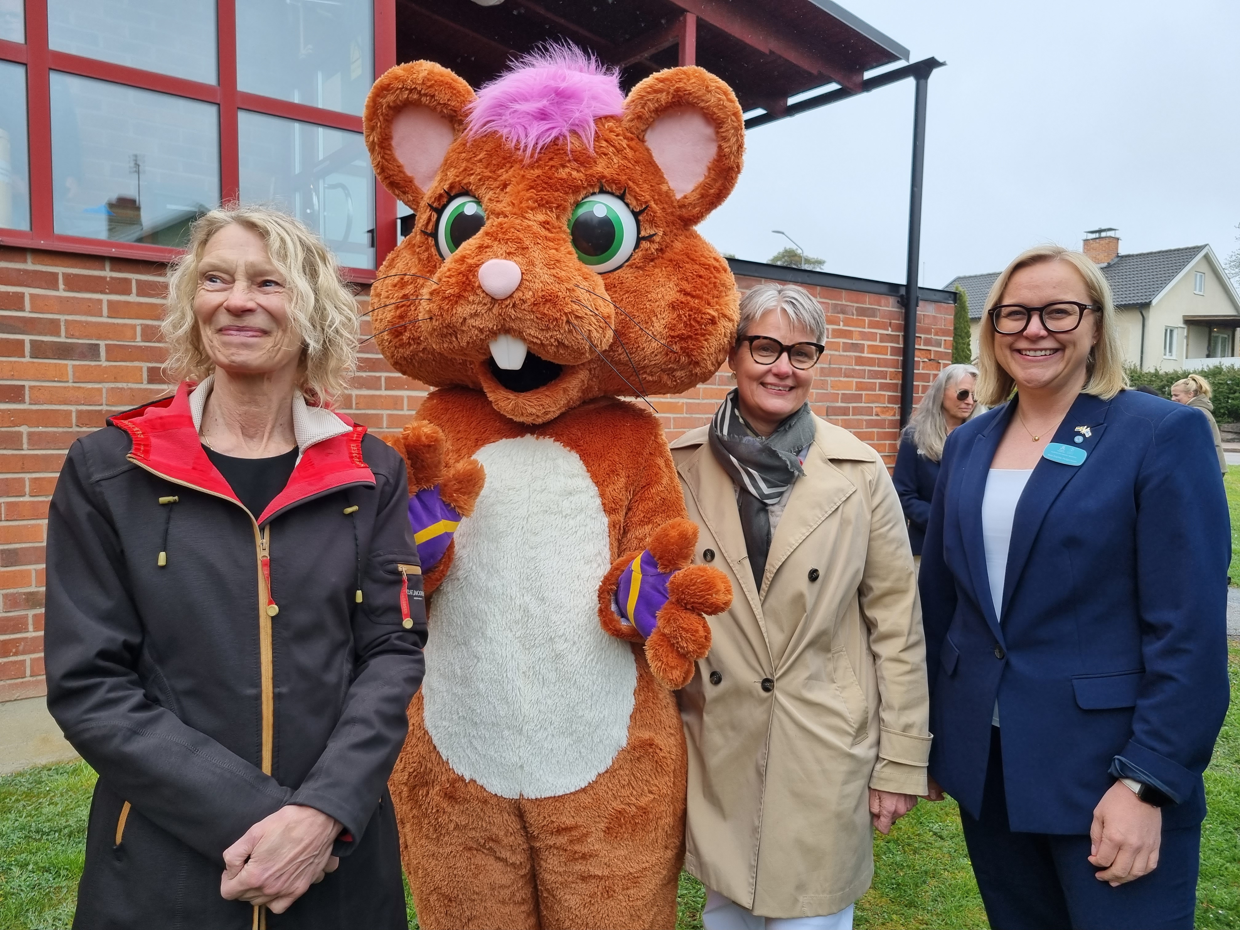 Tre personer står på rad tillsammans med stor gosedjursfigur. 