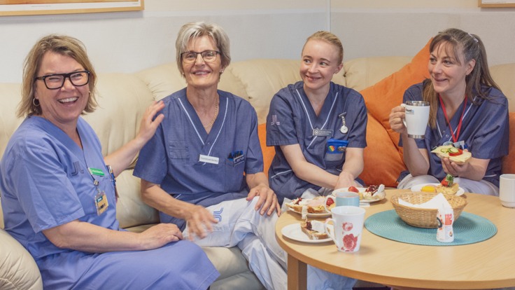 Fyra kvinnor i sjukvårdskläder sitter i en soffa och fikar och skrattar.
