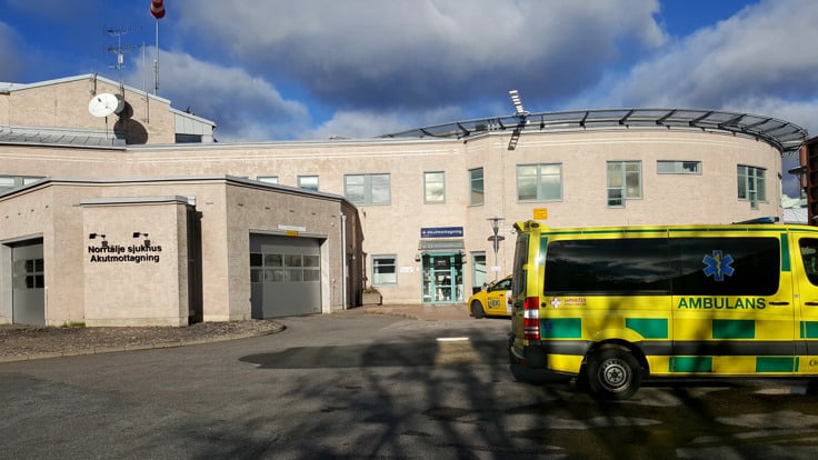 Ambulans framför tegelbyggnad. På en skylt på väggen står det Norrtälje sjukhus Akutmottagning. 