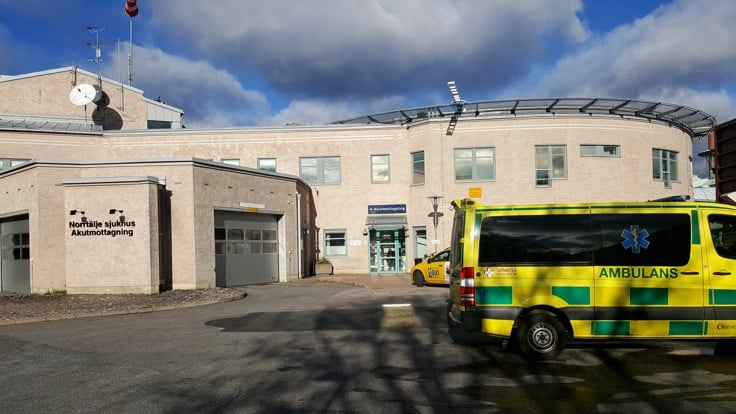 Ambulans framför tegelbyggnad. På en skylt på väggen står det Norrtälje sjukhus Akutmottagning. 