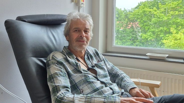 Patienten Anders tittar in i kameran sittandes på en stol framför ett fönster med grönskande träd utanför. 