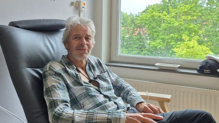 Patienten Anders tittar in i kameran sittandes på en stol framför ett fönster med grönskande träd utanför. 