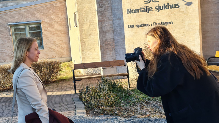 En kvinna fotograferar en annan kvinna framför en solbelyst vägg med texten Norrtälje sjukhus.