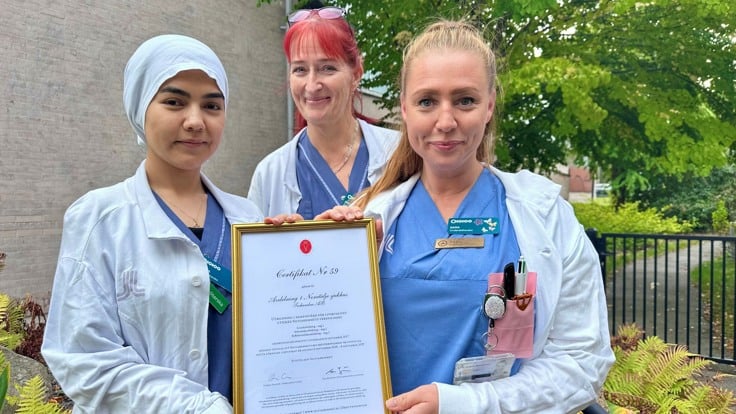 Tre personer klädda i vårdkläder står på rad utomhus. I bakgrunden syns en bit av en fasad och ett träd. Två av personerna håller i ett inramat papper.