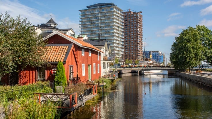 Rött trähus i förgrunden, vatten och två stora höghus