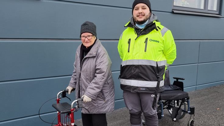 Vinterklädd kvinna med rullator står utomhus. Bredvid henne står en man klädd i reflexjacka.