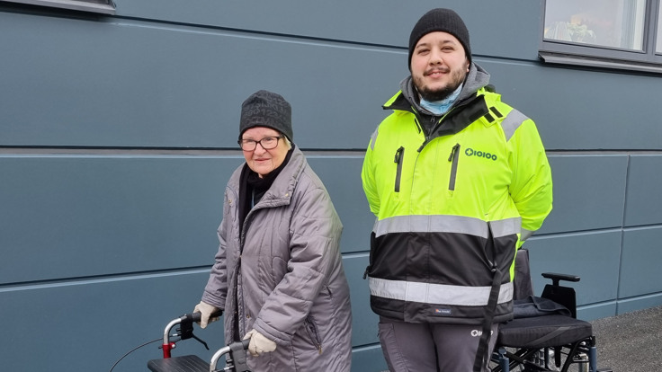 Vinterklädd kvinna med rullator står utomhus. Bredvid henne står en man klädd i reflexjacka.