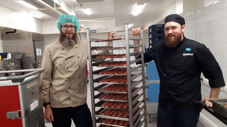 Två män i kockkläder står i storkök. Mellan sig har de en ställning med plåtar fulla med köttbullar.