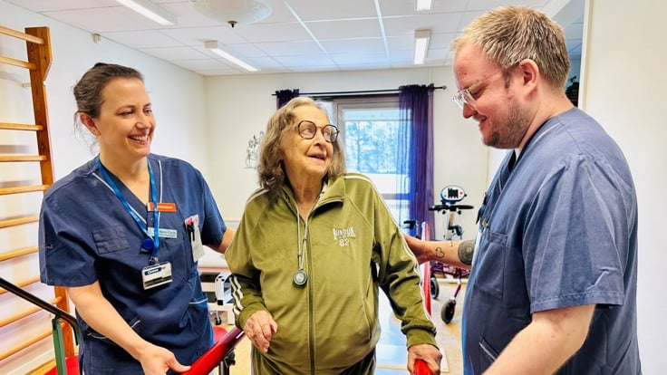Tre personer står i träningslokal, två är klädda i blå vårdkläder.