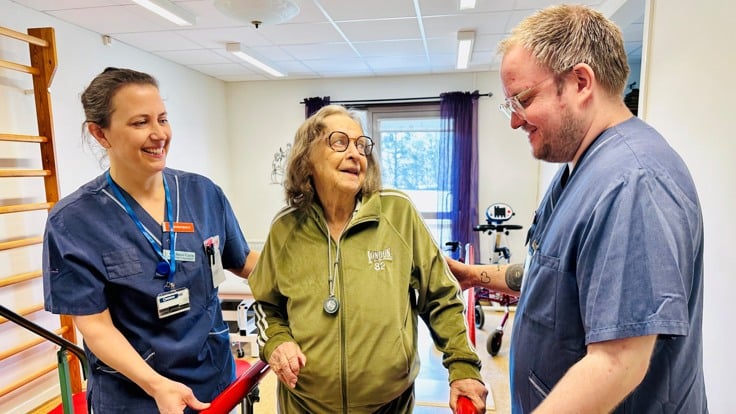 Tre personer står i träningslokal, två är klädda i blå vårdkläder.