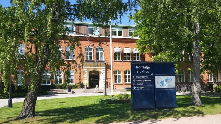 En äldre tegelbyggnad omgiven av grönska. I förgrunden en skylt med texten Norrtälje sjukhus.