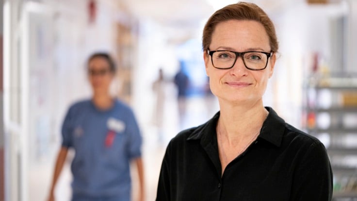 En kvinna med glasögon står i en korridor. I bakgrunden passerar en person.