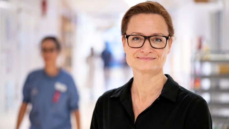 En kvinna med glasögon står i en korridor. I bakgrunden passerar en person.