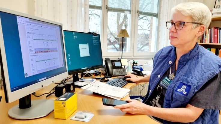 Kvinna sitter vid skrivbord med dator och håller en mobiltelefon i handen.