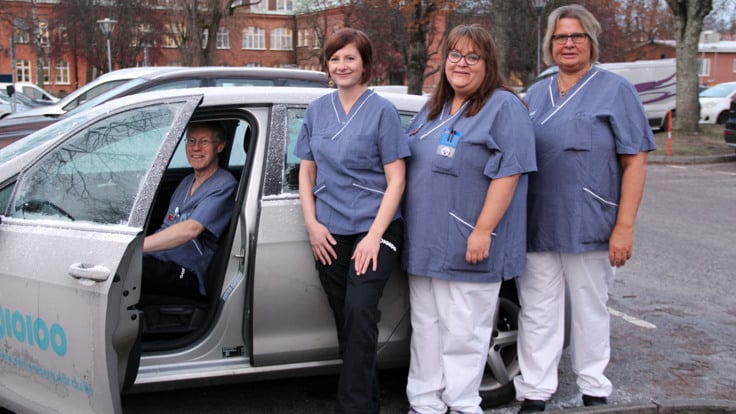 Tre kvinnor klädda i sjukvårdskläder står på rad vid bil. En man sitter på förarsätet i bilen . Bildörren är öppen.