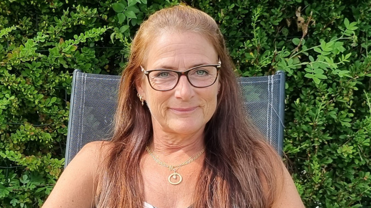 Leende kvinna med glasögon sitter i stol. Grönska i bakgrunden.
