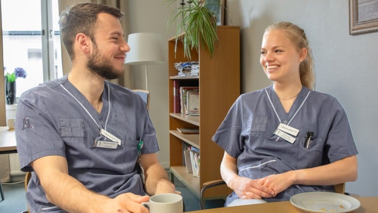 Två personer sitter vid bord och fikar. Ler mot varandra. De har namnbrickor med texten "läkarstudent". 