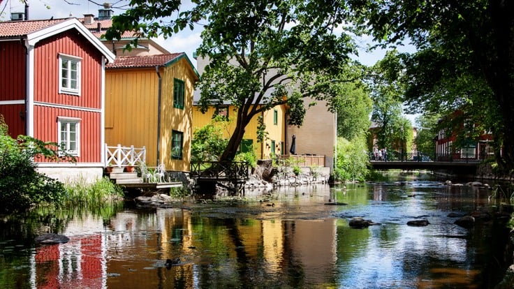 Norrtäljeån ligger spegelblank, till vänster i bilden syns pittoreska trähus i rött och gult, Ett träd sträcker sig ut över vattnet.
