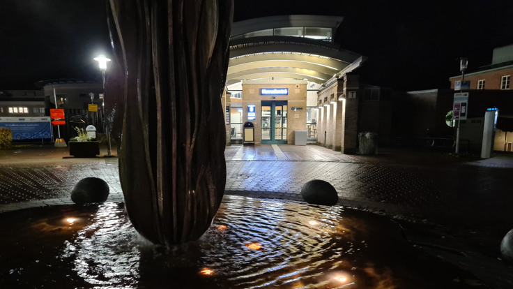 Stor byggnad med upplyst entré. I förgrunden en fontän. Det är mörkt ute.