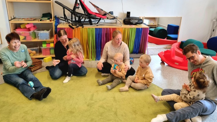 Vuxna och barn sitter på en matta på golvet i förskolemiljö.
