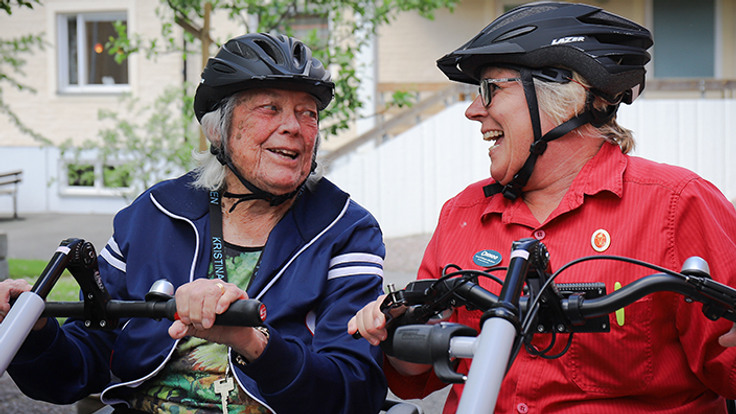 Två äldre skrattande kvinnor på en parcykel. 