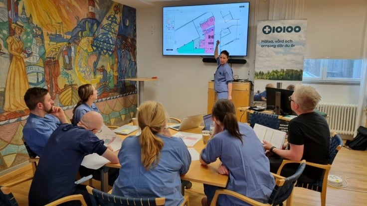 Grupp av människor sitter runt ett bord med papper på. En person står upp och pekar på en tv-skärm. 