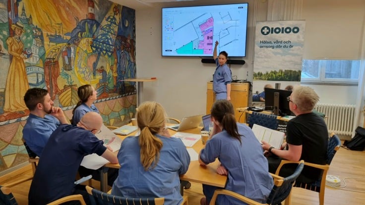 Grupp av människor sitter runt ett bord med papper på. En person står upp och pekar på en tv-skärm. 