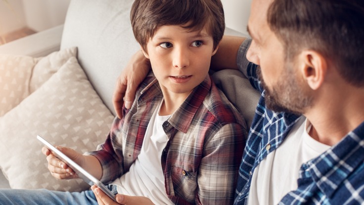 Man och pojke sitter i en soffa och tittar på varandra. Mannen håller armen om pojken.