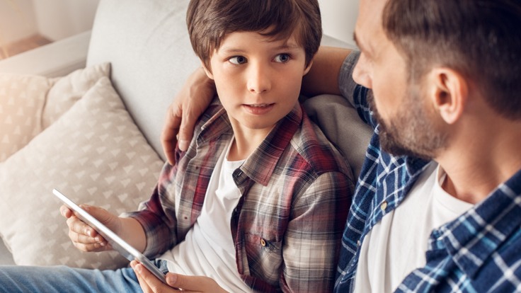 Man och pojke sitter i en soffa och tittar på varandra. Mannen håller armen om pojken.