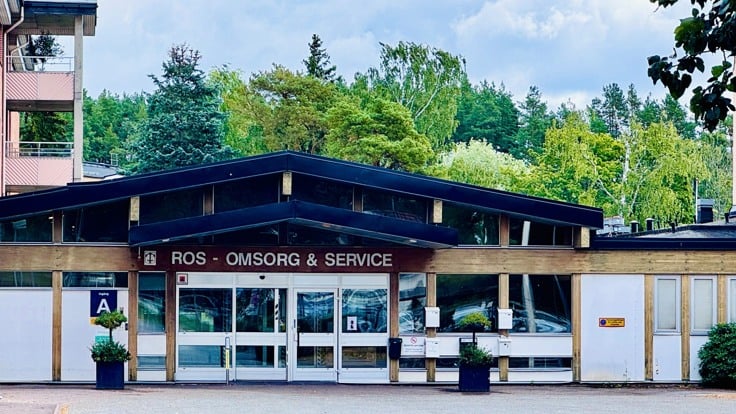 Byggnad med träd i bakgrunden. På fasaden står det ROS Omsorg & Service.