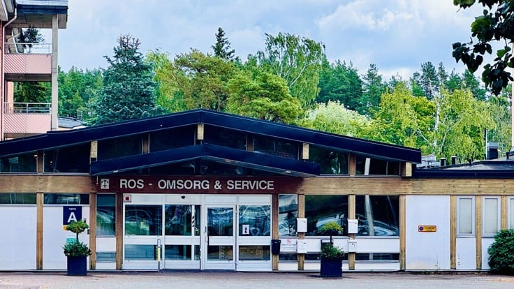 Byggnad med träd i bakgrunden. På fasaden står det ROS Omsorg & Service.