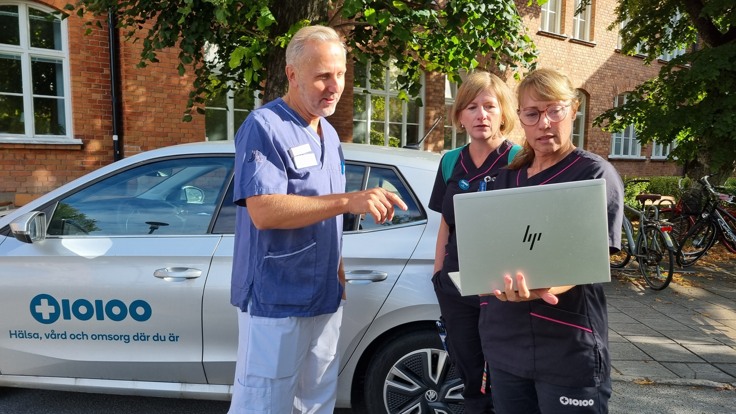 Tre personer i sjukvårdskläder står framför tegelbyggnad och bil och tittar på en bärbar dator.