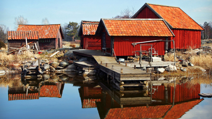 Röda sjöbodar står i grupp. en brygga sträcker ut sig mot spegelblankt vatten.