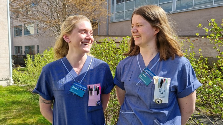 Två kvinnor i blå vårdkläder ler mot varandra.