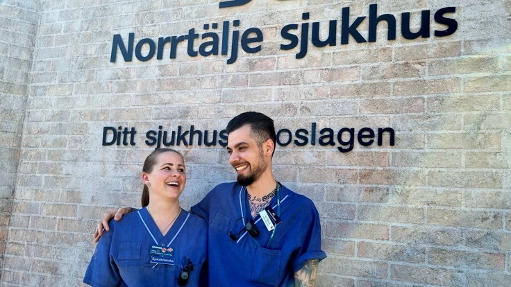 Två personer står och ler och håller om varandra framför en tegelvägg med texten Norrtälje sjukhus.