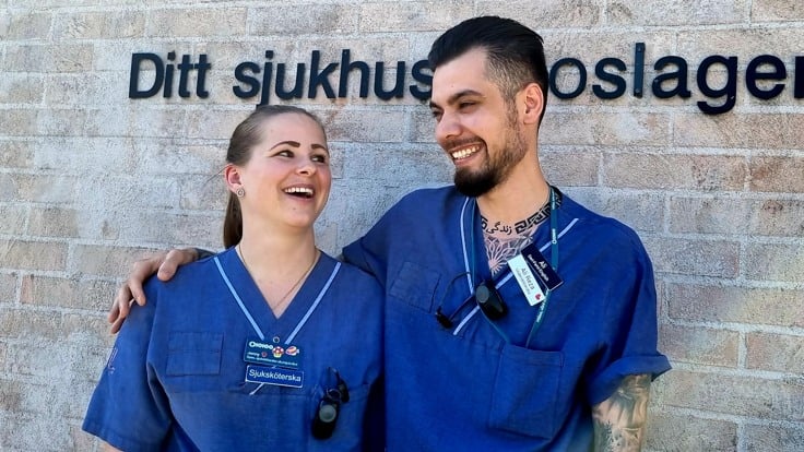 Två personer står och ler och håller om varandra framför en tegelvägg med texten Norrtälje sjukhus.