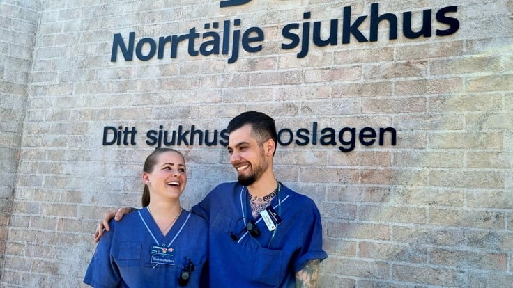Två personer står och ler och håller om varandra framför en tegelvägg med texten Norrtälje sjukhus.
