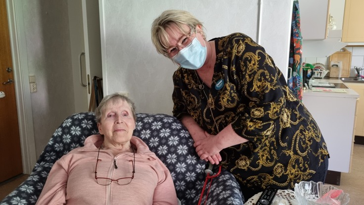 Äldre kvinna sitter i fåtölj, yngre kvinna står bredvid, tittar in i kameran och ler.