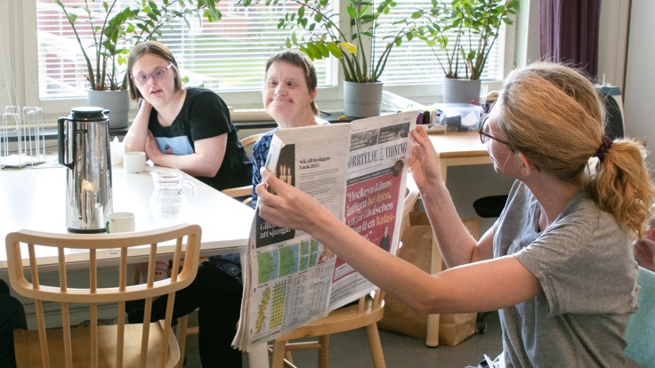 Personal håller upp en dagstidning och visar deltagare som sitter vid ett bord