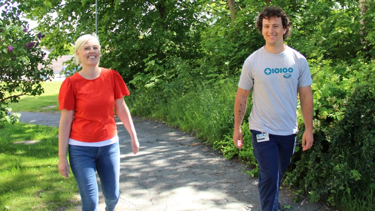 En man och en kvinna går på en gångväg i sommargrönska. De ler och tittar i kameran. Mannen har t-shirt med Tiohundras logga.